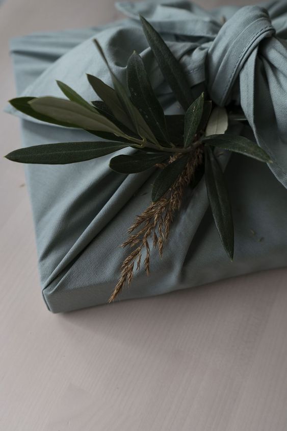 Foto de um detalhe de uma embalagem de pano, feita de um tecido azul claro. Parece ser um livro. Em cima da amarração tem um galhinho de uma planta.