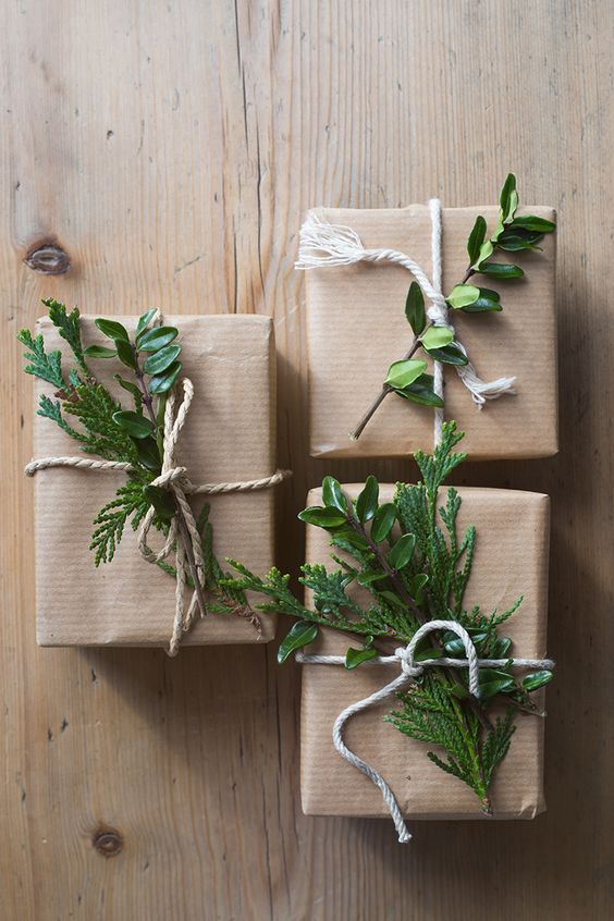 Três pacotes de presente embrulhados com papel pardo, barbante e galhinhos de plantas presos no barbante. Eles estão em cima de o que parece ser uma mesa de madeira.