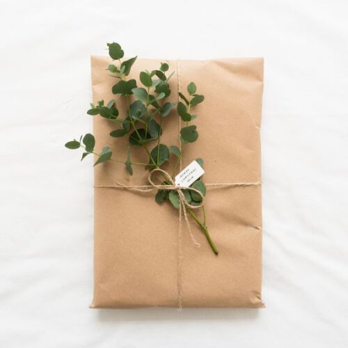 Um pacote de presente de papel pardo com um barbante amarrando e um galhinho verde de alguma planta. A foto é de cima e o pacote está em cima de um tecido branco.