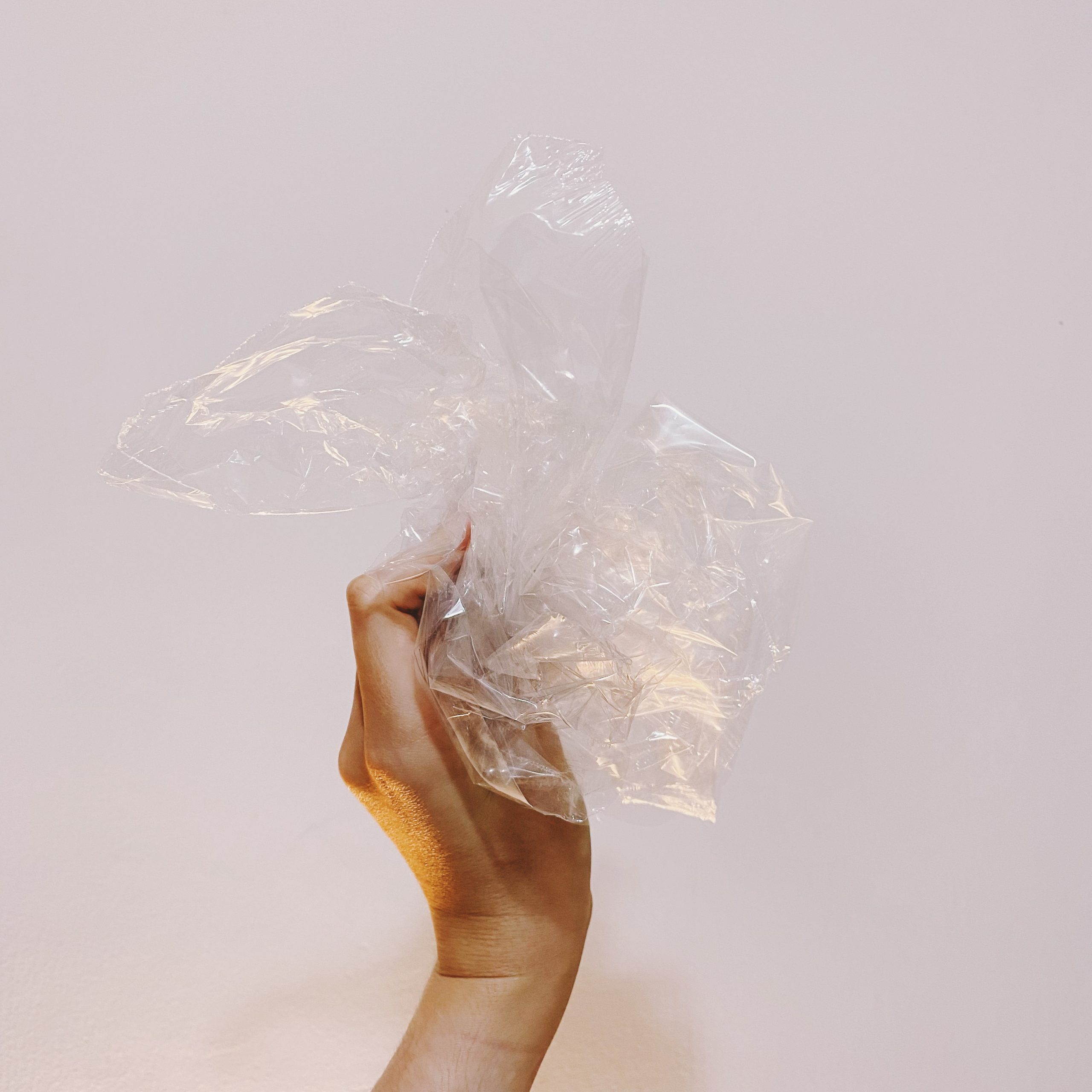 Fotografia, de dia, em ambiente interno de uma mão de uma pessoa branca segurando um monte de plástico transparente, do tipo fininho, na frente de uma parede branca